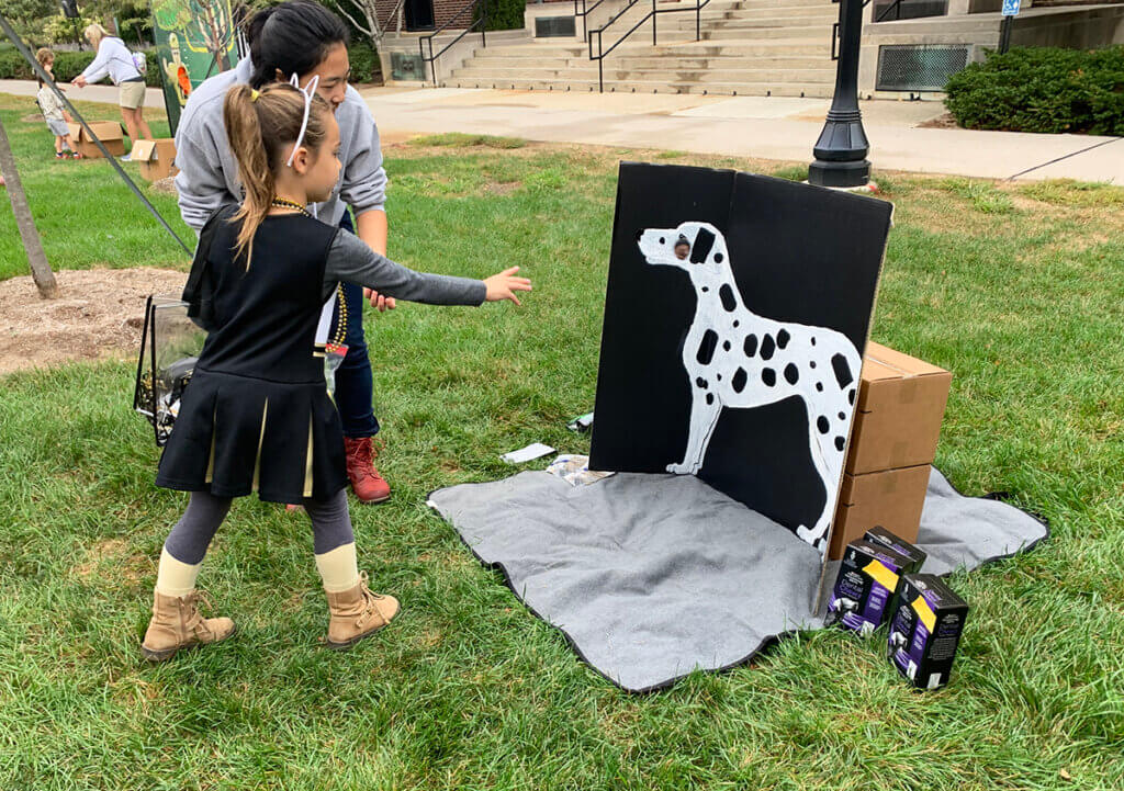 A little girl wearing a Purdue cheerleading outfit in black and gold tosses a "tick" onto the spotted dog backdrop as a PVM volunteer stands by to assist