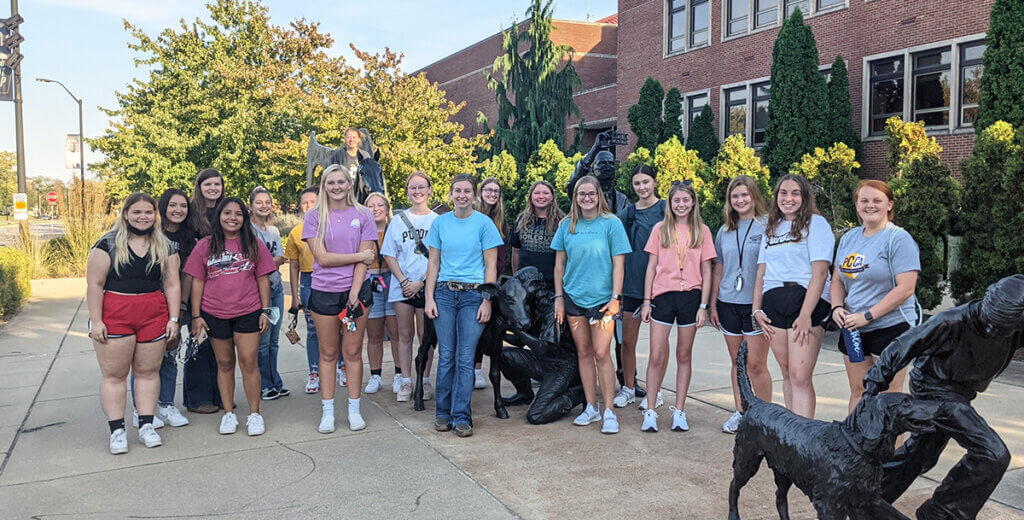 A group of female students join together smiling standing in line with the Continuum sculpture in front of Lynn Hall