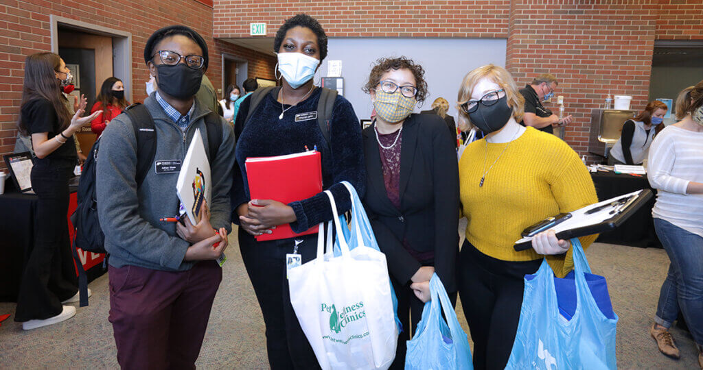 A group of four students stand together wearing facemasks and holding armfuls of giveaways