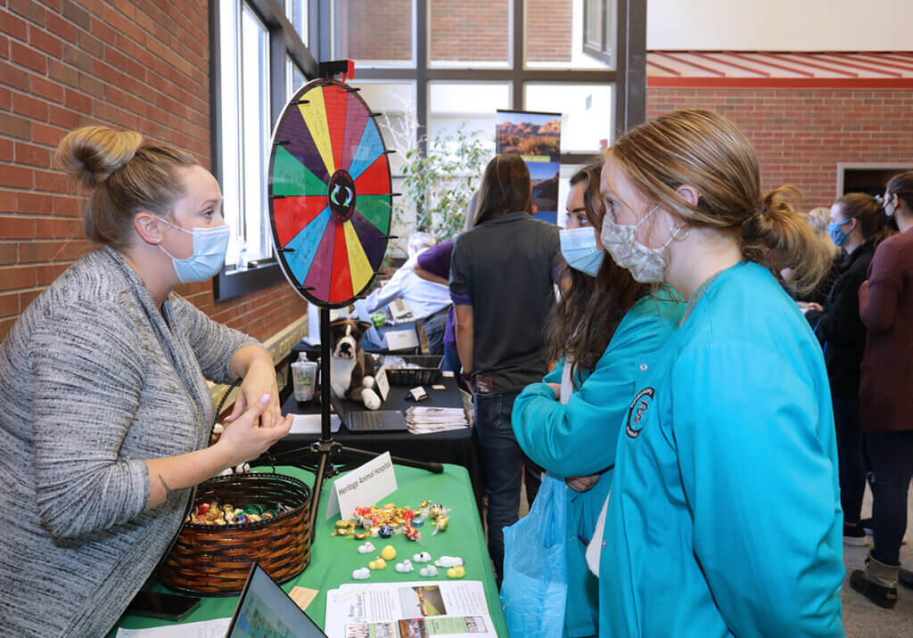 A representative stands next to a prize wheel speaking with veterinary nursing students