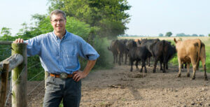 A man stands by a fence with cows walking in the background on a farm.