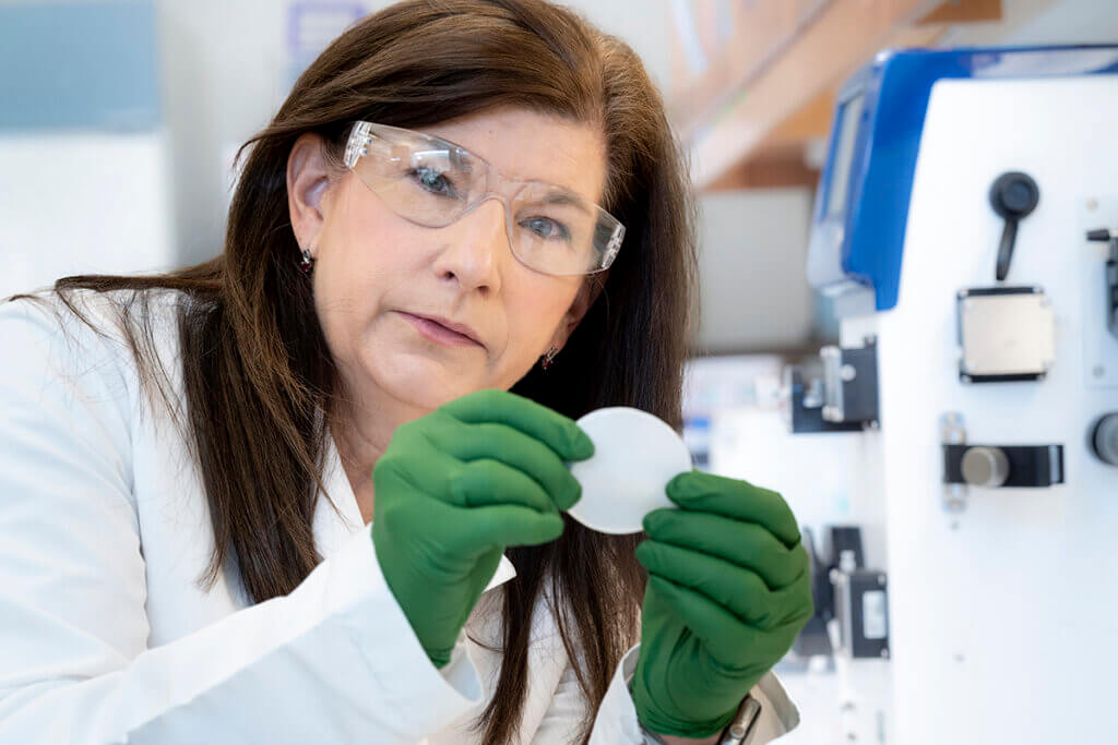 Sherry, wearing a lab cat, safety glasses, and gloves, holds up a collagen sheet