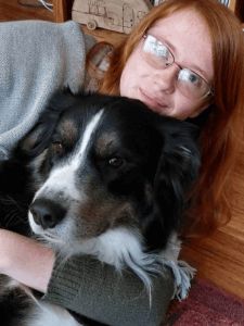 A person with red hair and glasses is cuddling a large black and white dog.