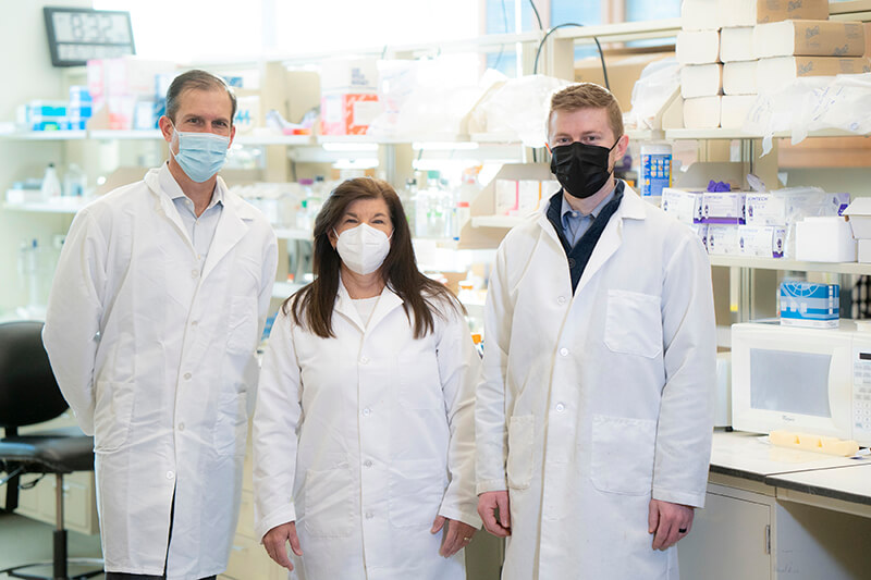Andy, Sherry, and TJ stand together wearing masks and lab coats with the lab in the background