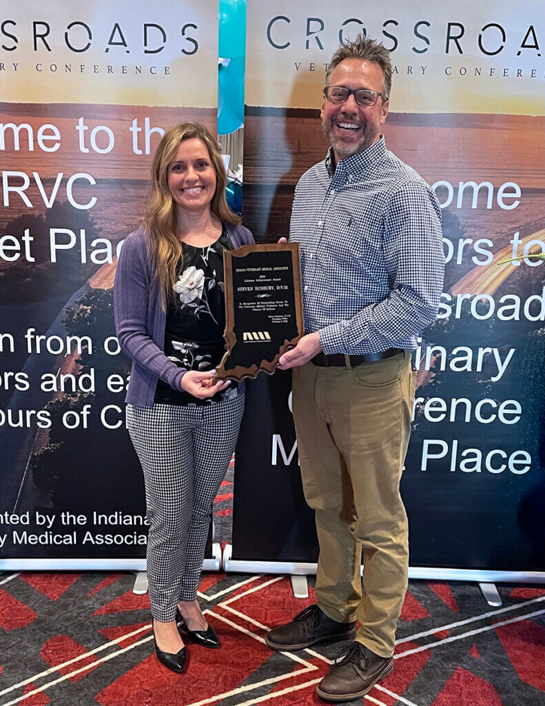 Dr. Christner and Dr. Sunbury stand together holding his plaque against a banner backdrop