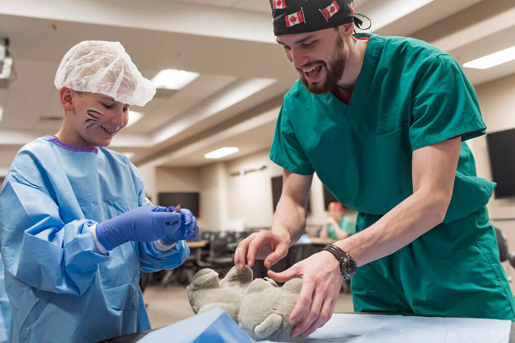 A child and an adult wearing medical scrubs and gloves engage in a hands-on learning activity involving a stuffed animal.