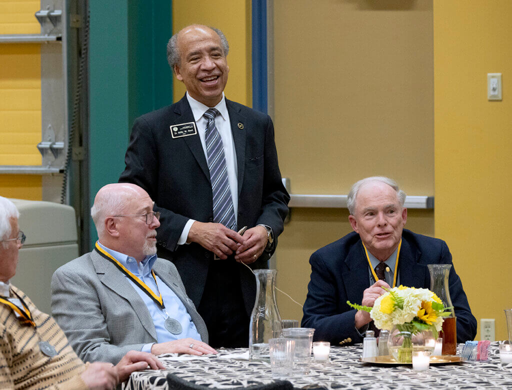 Dr. Kinnard sits beside classmates at a table as Dean Reed stands beside him smiling