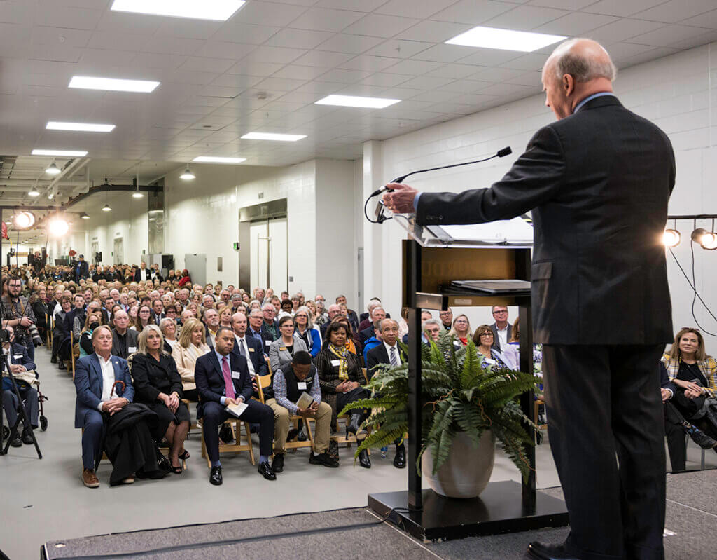 President Daniels stands on stage with the crowd of seated and standing guests shown in front of him