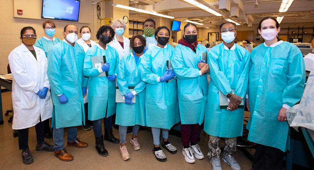UAPB students and mentors, wearing lab coats and smocks and face masks, gather for a group photo in a lab space