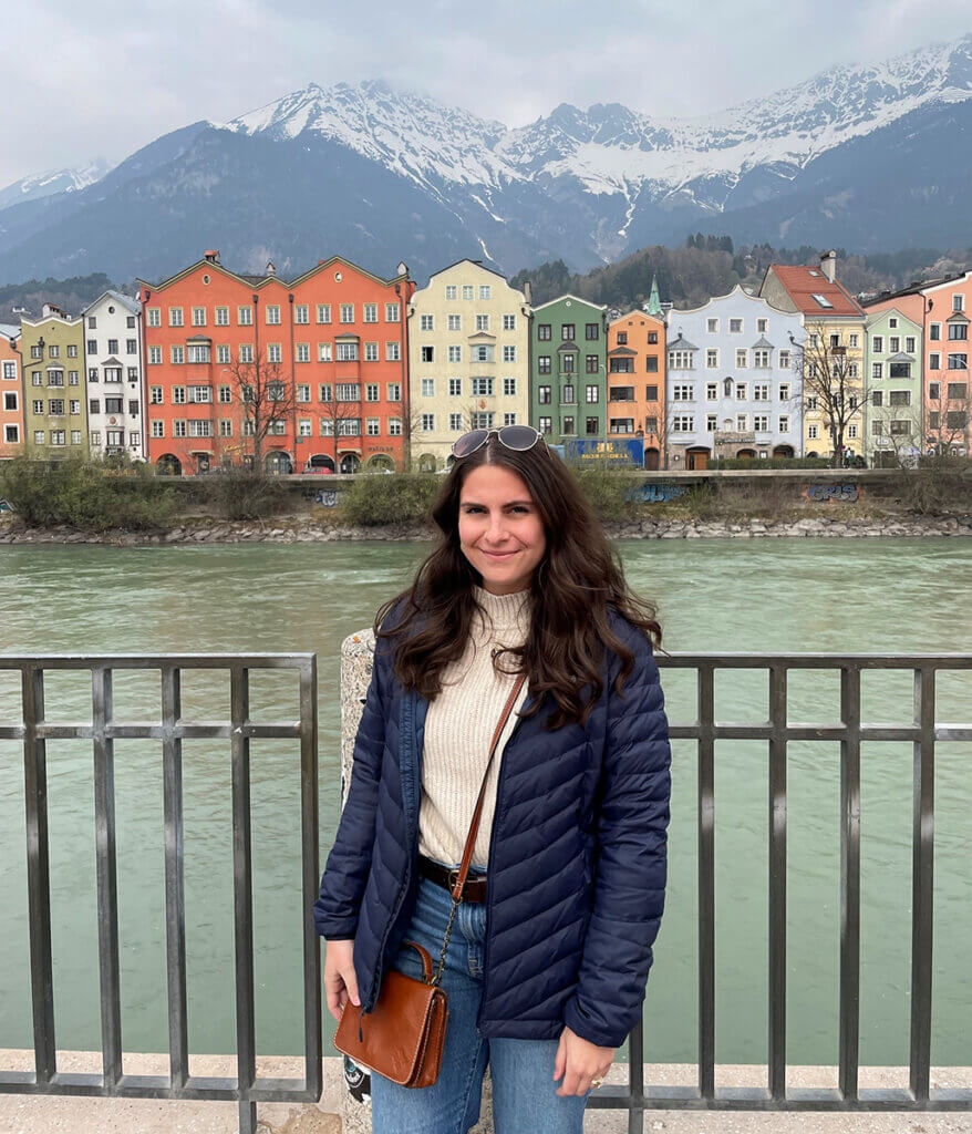 Katelynn stands smiling with a river, colorful buildings, and mountains in the background