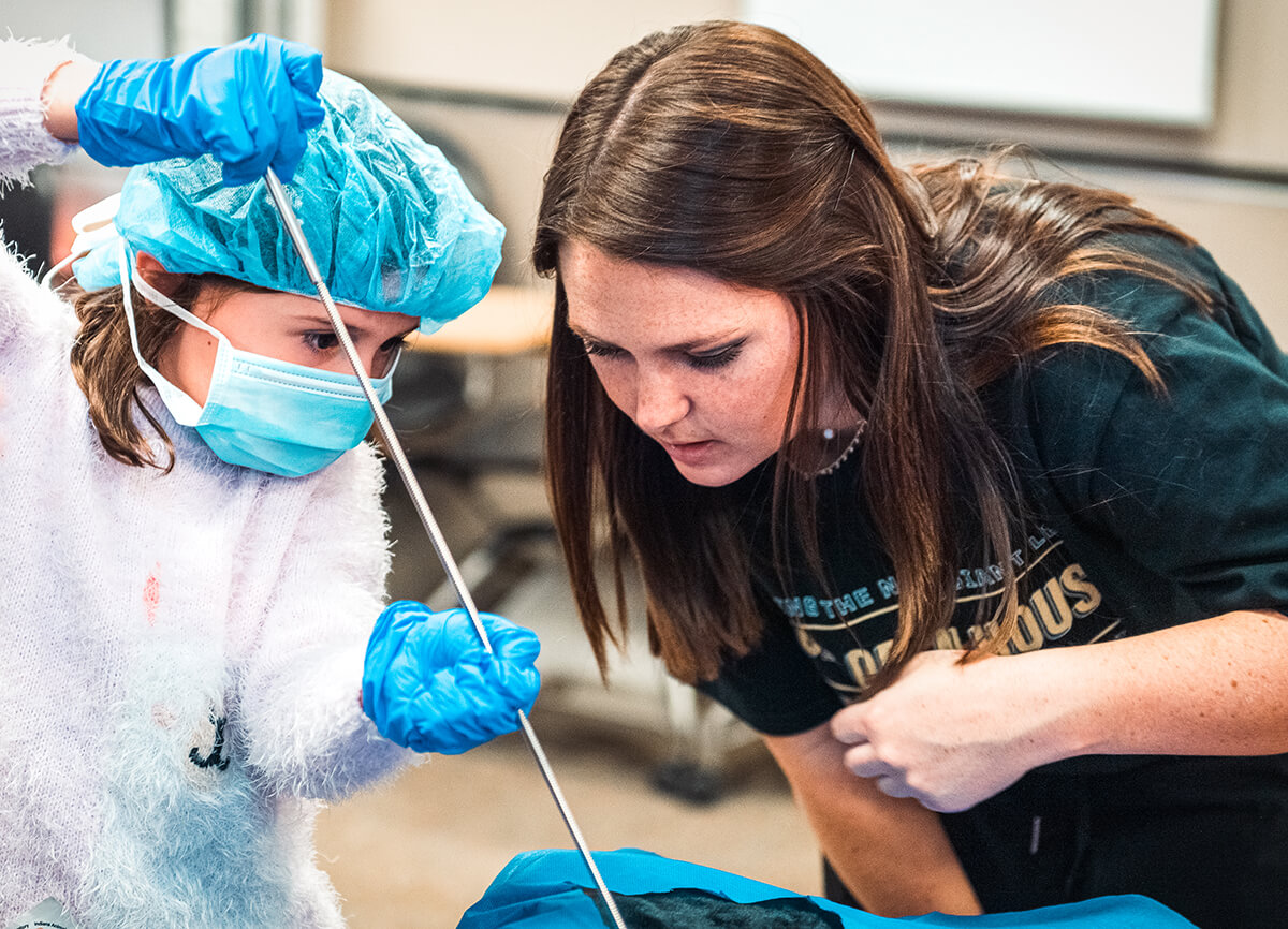 A young girl wears a surgical hat, mask, and gloves as she and a PVM student intently look at the task at hand in the Teddy Bear Clinic