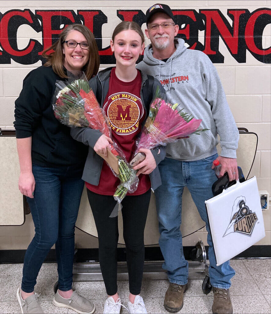 Brittany, her daughter, and husband smile into the camera as her daughter holds bouquets of flowers