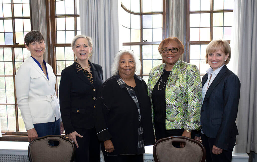 The women stand together smiling with the sun brightening the window behind them