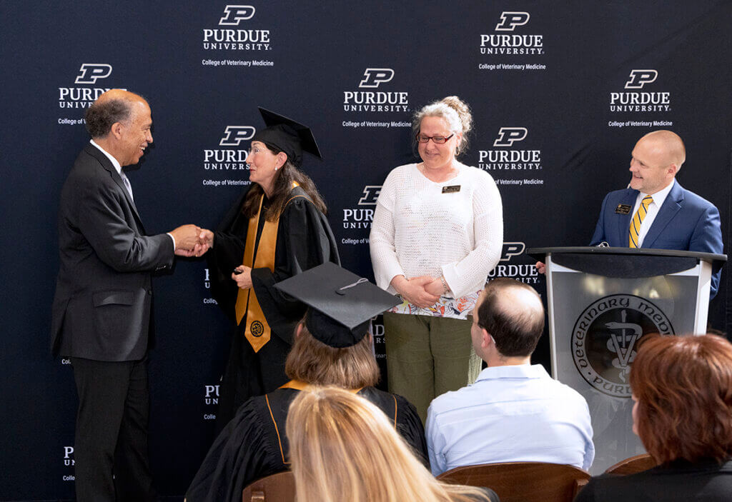 Dean Reed shakes Brenda's hand alongside Holly and Dr. Brown as the audience looks on. Brenda is wearing her graduation cap and gown.