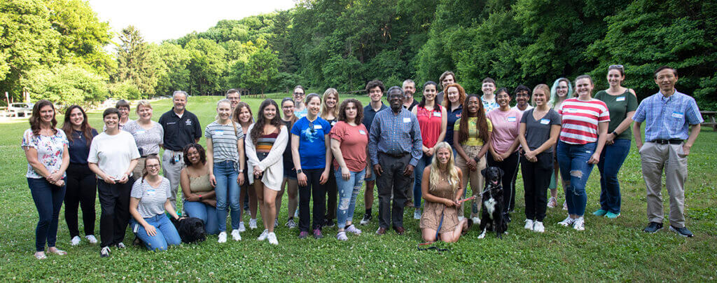 Picnic attendees gather for a group photo including some pups with a dense green treeline in the background