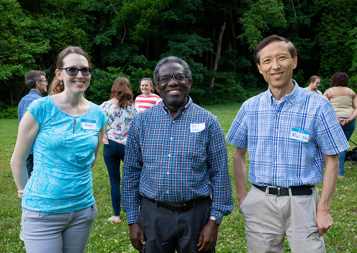 Amanda, Eli, and Riyi stand together smiling at the camera as attendees talk in groups in the background