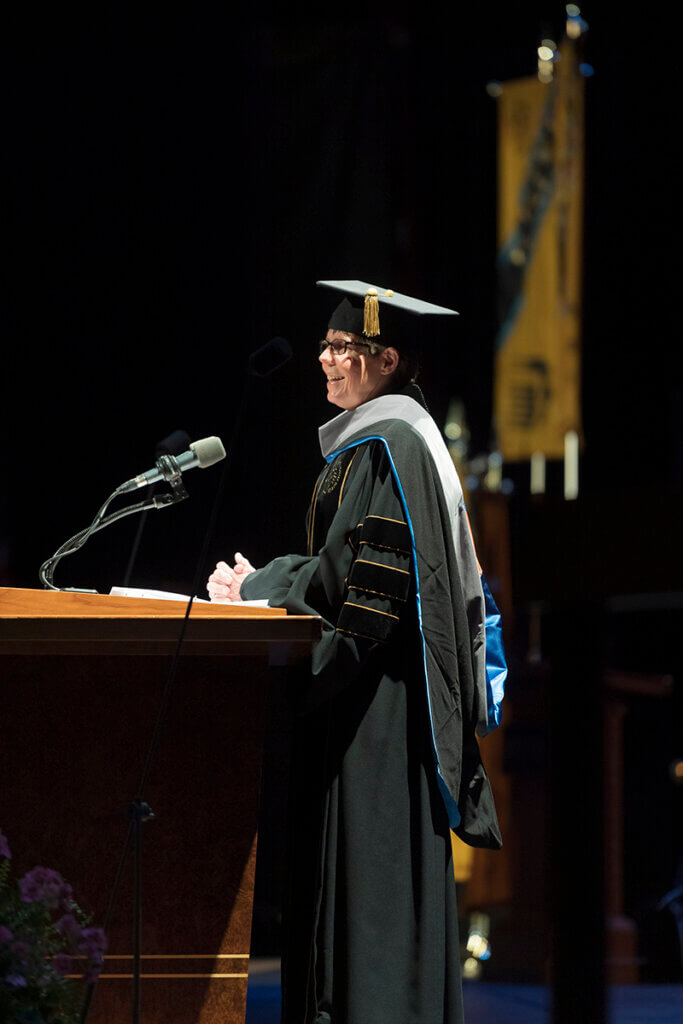 Dr. Knapp smiles as she speaks from a podium on the stage wearing traditional commencement garb