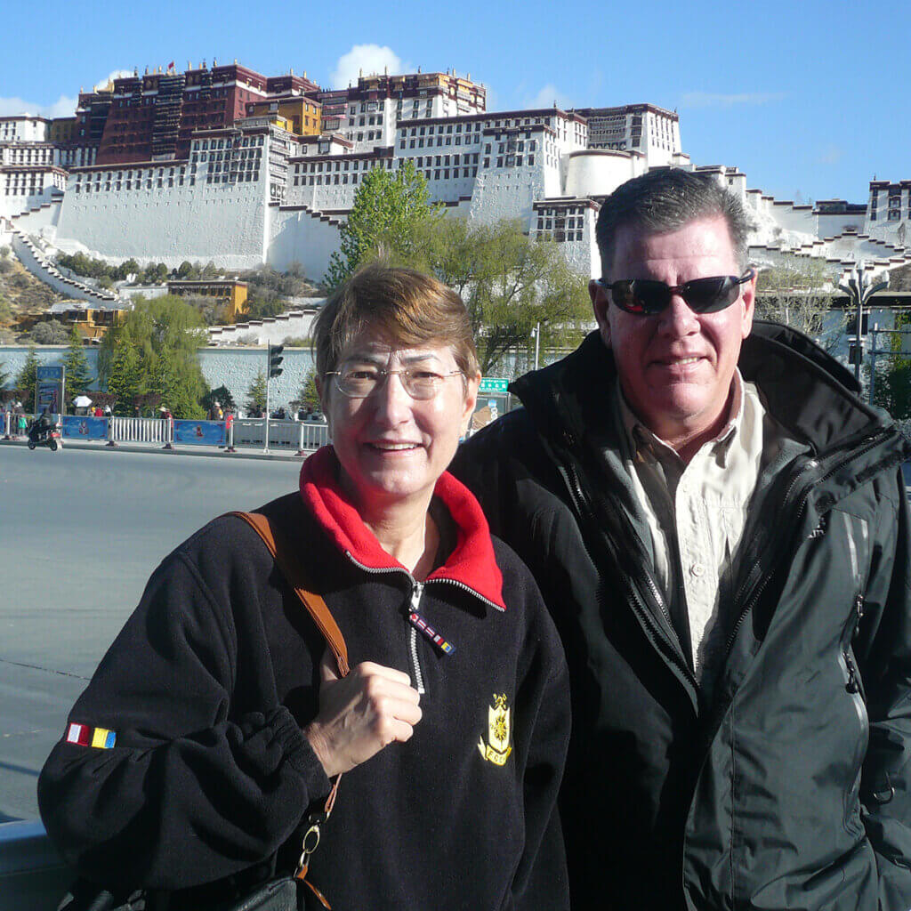 Brenda and her husband stand together smiling with the Potala Palace in the background atop a hill