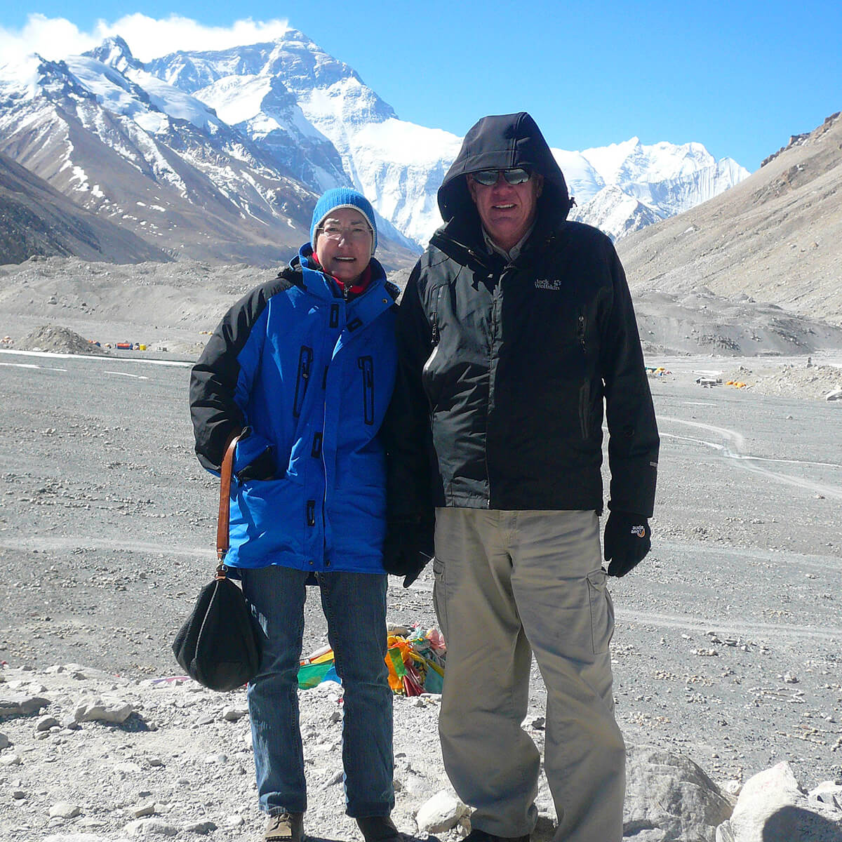 Brenda and John stand together smiling wearing winter clothes on rocky terrain with the snow caps of Mount Everest in the background