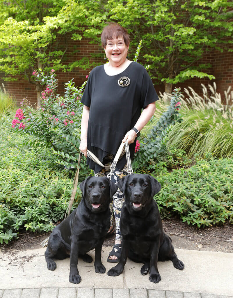 Joanne stands smiling as her dogs, two black Labs, sit nicely in front of her against a background of lush plants