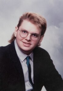 A portrait of a young man with glasses and a mullet hairstyle, wearing a suit and tie against a soft, neutral background.
