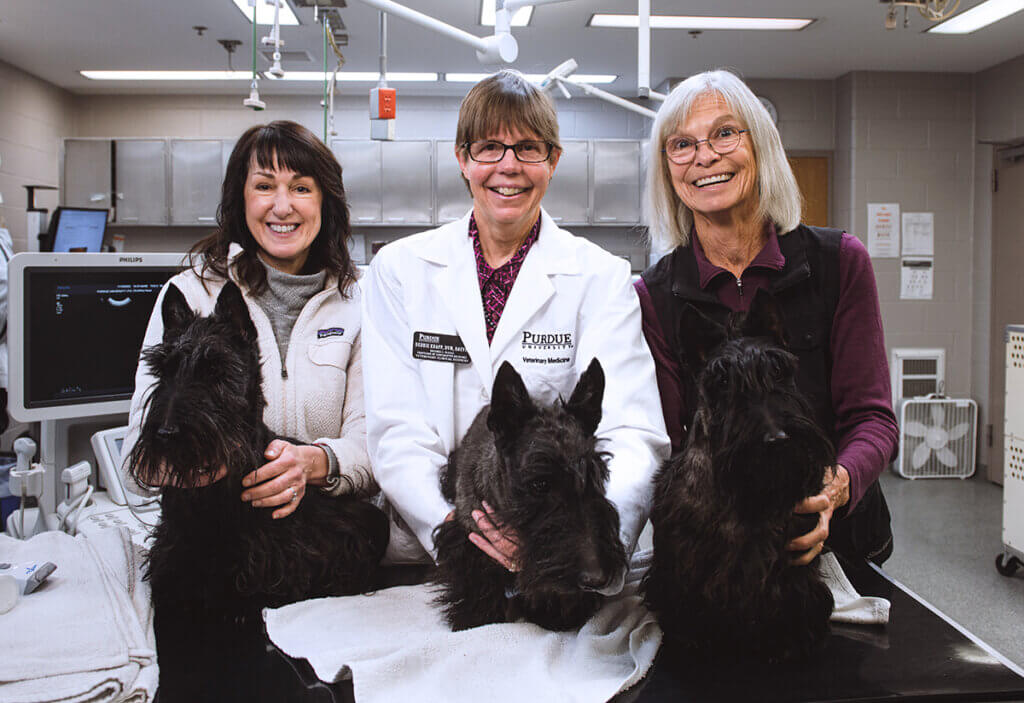 Dr. Knapp wears a white coat and stands in between two women all smiling holding black Scottish terriers in the oncology suite