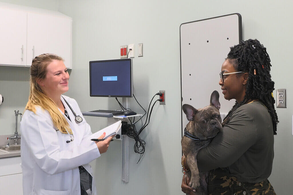 Veterinary student wearing a white coat speaks with Angel as she holds her pup in an exam room