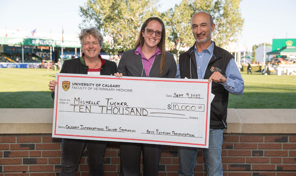 The trio stand outside against a short brick wall smiling and holding up an oversized check