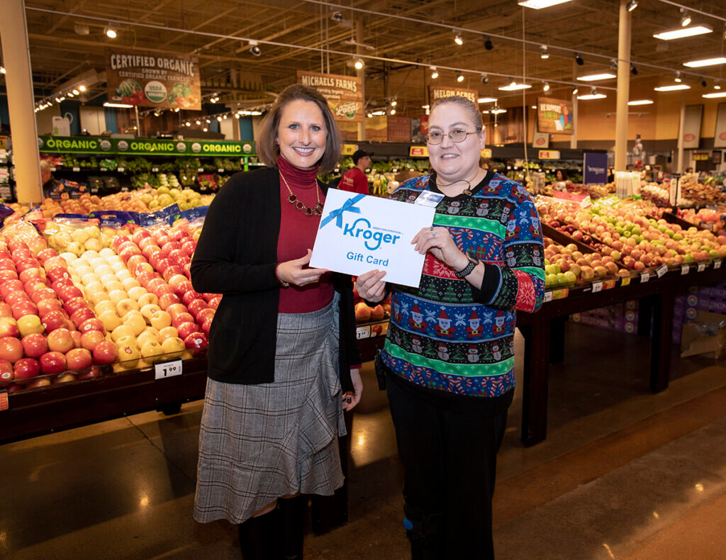 Amanda and Jennifer both smile as they hold up an oversized gift card in front of the produce section of the grocery store