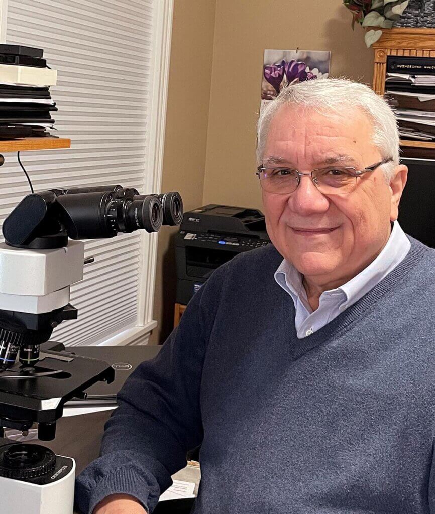Dr. DeNicola turns toward the camera smiling as he sits next to a microscope in his office
