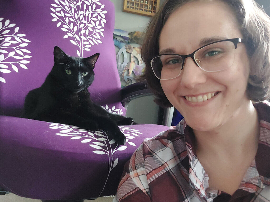 Megan smiles next to a black cat perched on a vibrant purple chair