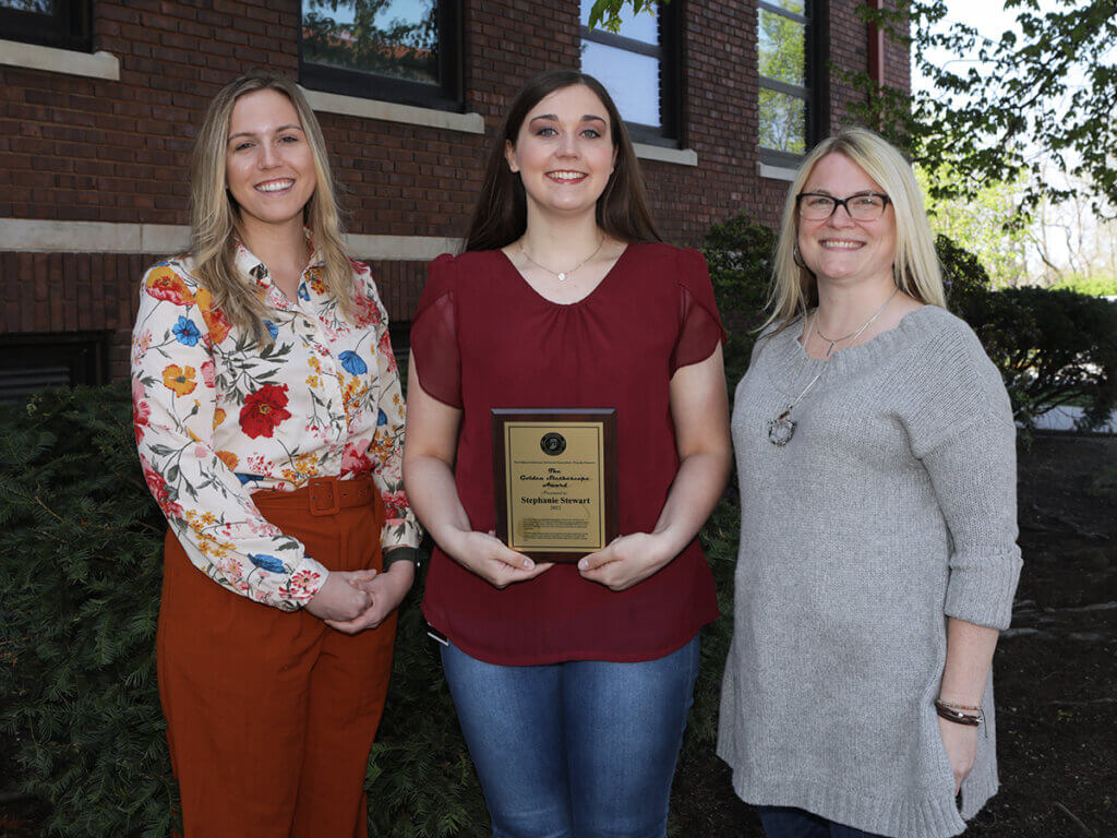 Stephanie smiles holding her award plaque with Crystal and Myranda standing on either side of her outside the VPTH building on campus