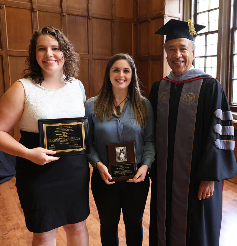 Patience and Miriam smile holding their award plaques as Dean Reed, wearing commencement robes, joins them following the college's graduation ceremony