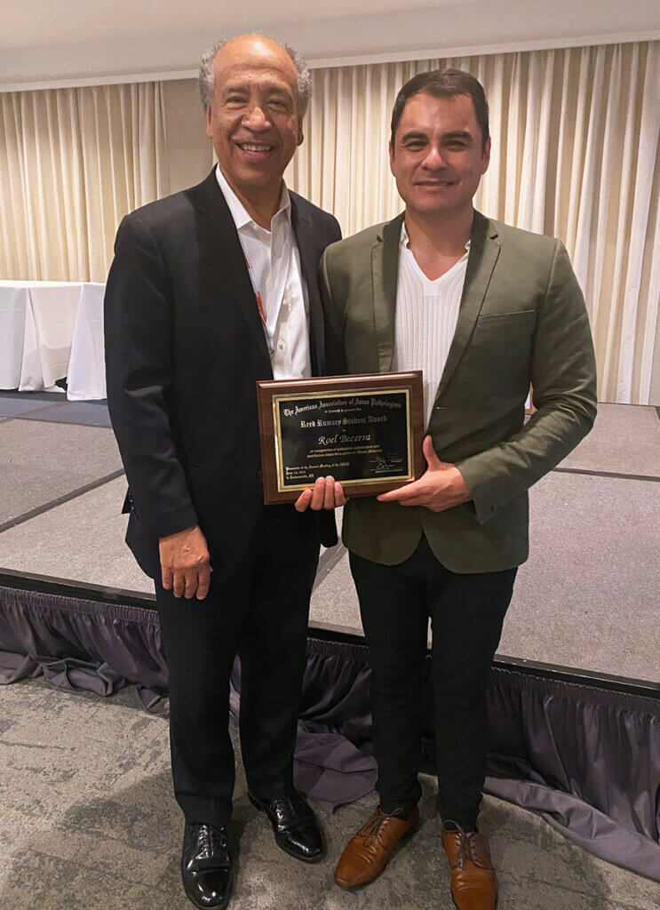 Dean Reed and Dr. Becerra stand together smiling in front of the stage as Dr. Becerra holds up his award plaque