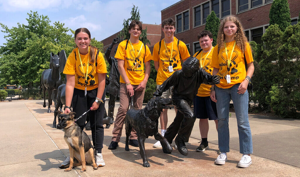 A group of five senior campers join together for a group photo with their camp dog at the Continuum sculpture in front of Lynn Hall