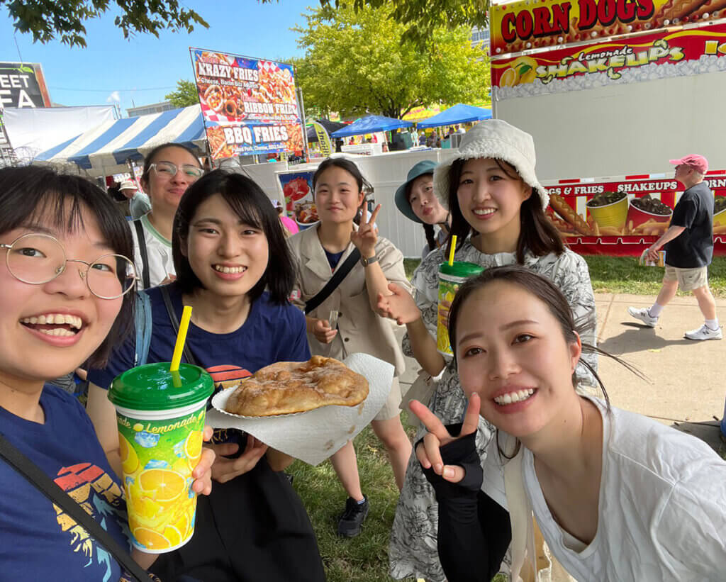The students flash peace signs and smile as they take a selfie at the State Fair with food and drink in hand.