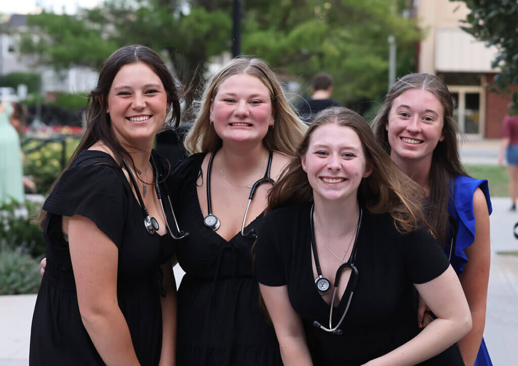 The four students stand close together smiling at the camera, wearing their stethoscopes around their necks.