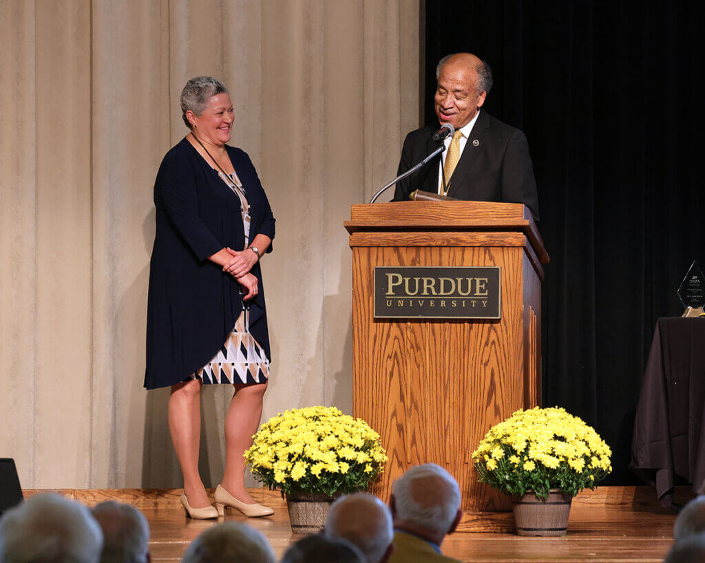 Pam smiles standing next to the dean as he shares some remarks on stage in recognition of her award.