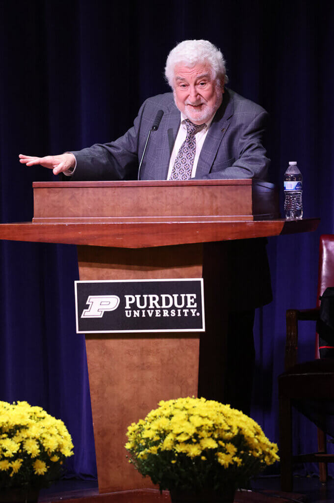 Dr. Beck stands behind a Purdue lectern in Stewart Center's Fowler Hall with bright yellow mums decorating the stage in front.