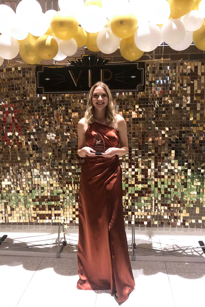 Madeline stands against a sparkly photo backdrop holding her award at the award gala