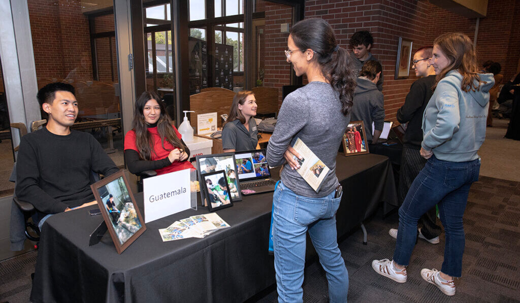 Students share their study abroad experiences with fellow students as they sit behind a table displaying photos and information on their experiences.