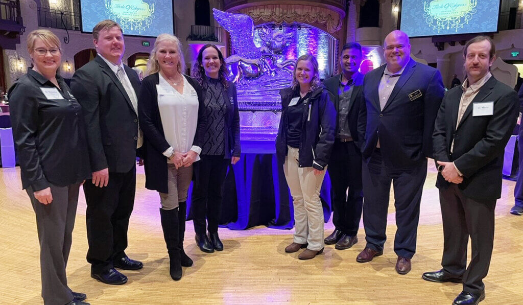 College faculty and staff in attendance join together for a group photo in front of an elaborate flying pig ice sculpture in the middle of the ballroom