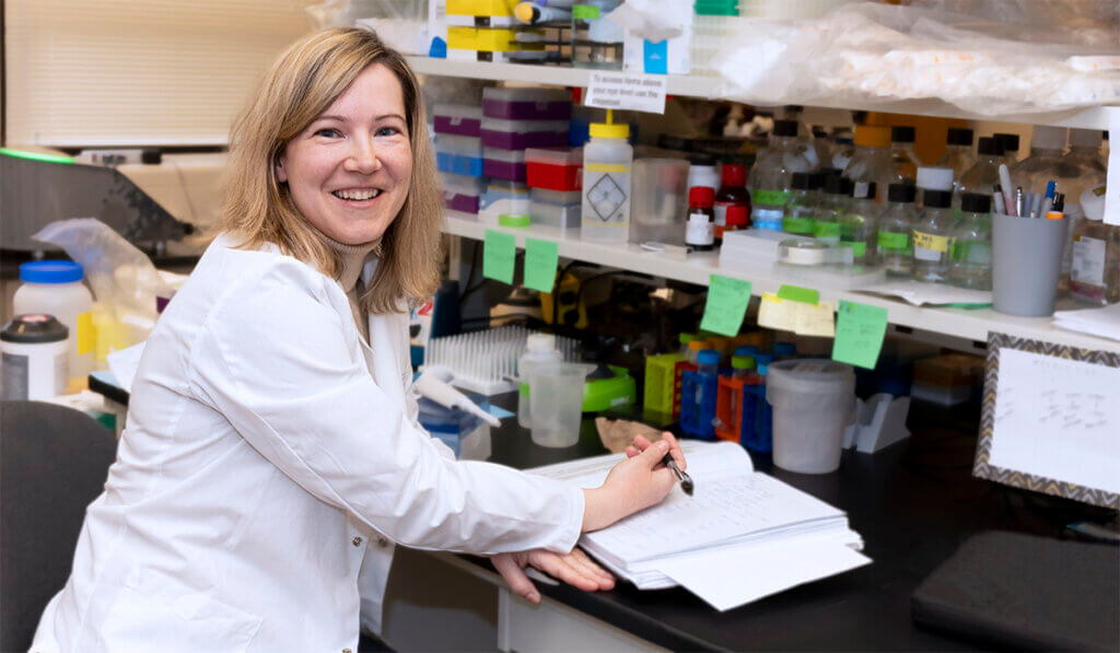Dr. Fortin pauses writing at a work station in her lab to smile at the camera