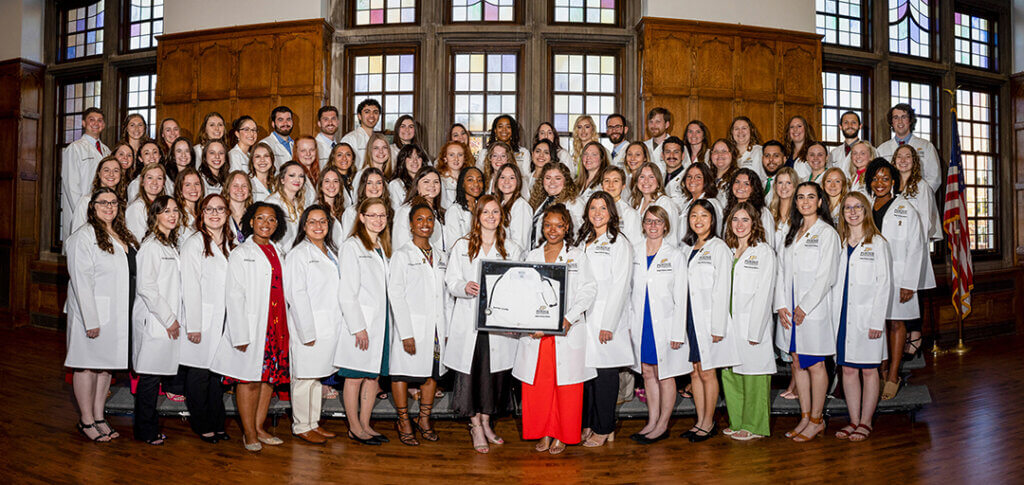 Wearing their white coats and holding the framed white coat of their classmate Aaron Lewis in his memory, members of the Class of 2025 gathered for their class photo.