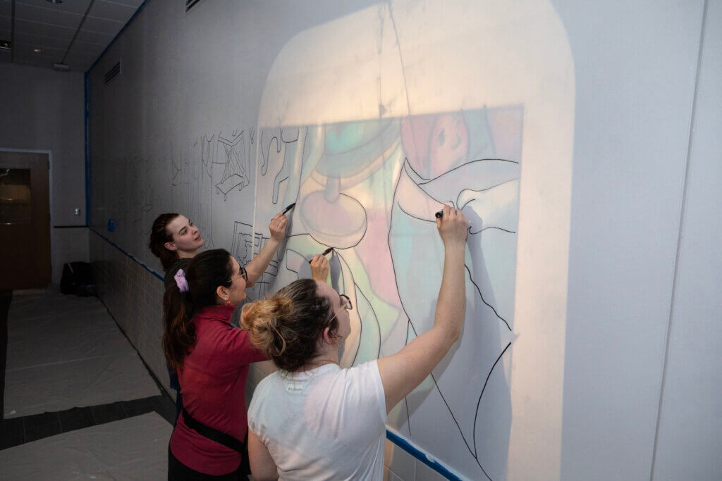 Once approved by the university, Tia’s mural design was outlined on the wall by volunteers who traced images of the drawing that were cast onto the blank surface with overhead projectors.