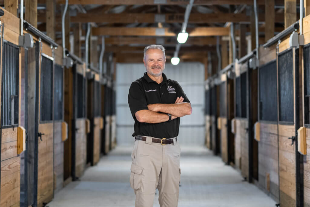 Man standing in a hallway with horse stalls on both sides of him. He is smiling with his arms crossed.