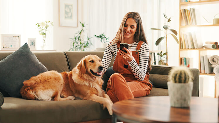 woman smiling, looking at her phone while petting a dog that is sitting next to her on a sofa