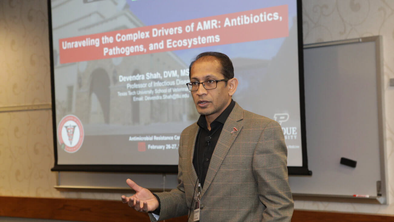 person wearing a tan suit jacket is giving a presentation, with information behind him on a projector screen