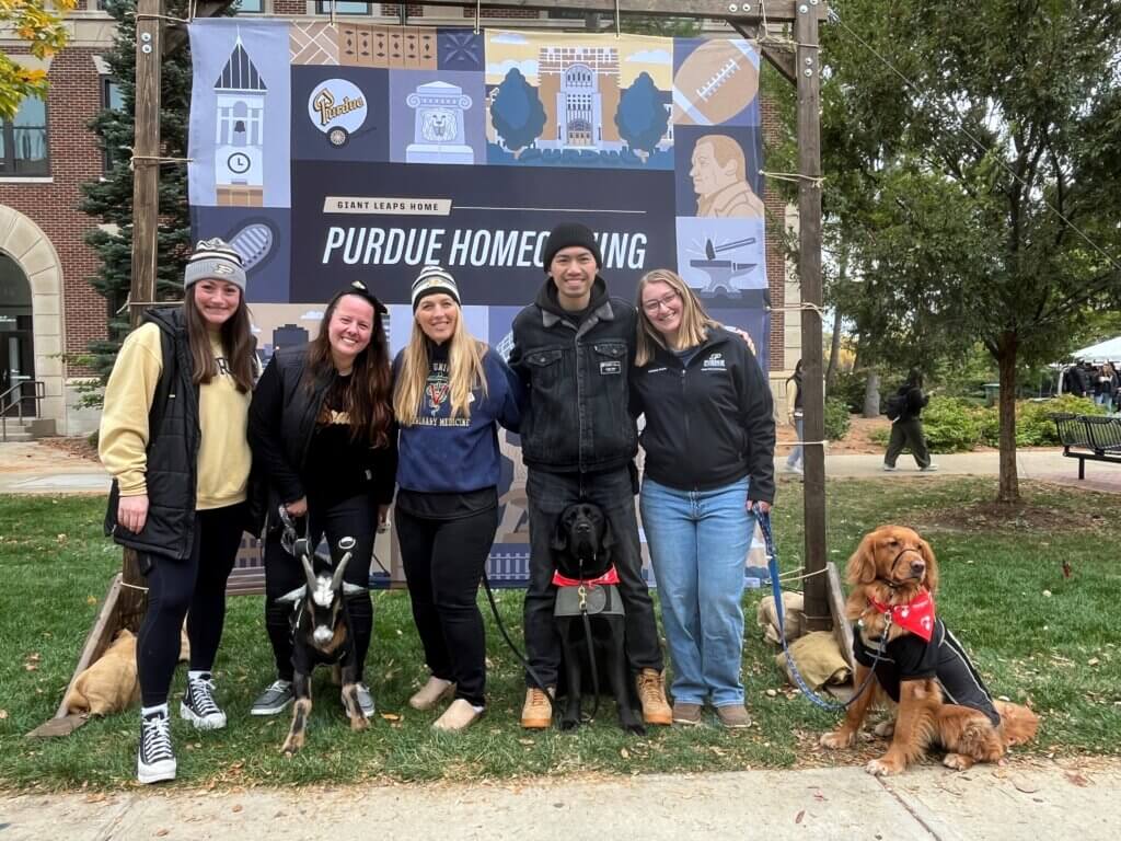 A group of people are standing outside in front of a large banner. The people are dressed for cold weather. There are 2 dogs sitting in front of the people and a goat standing in front of one person.