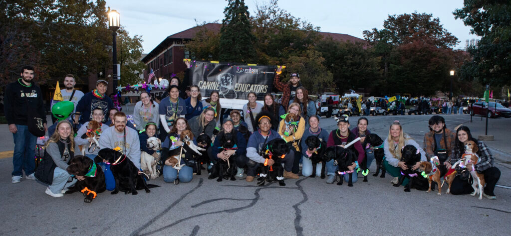 A large group of students are posing for an outdoor photo with several dogs. There is a banner behind the group that says "Canine Educators."